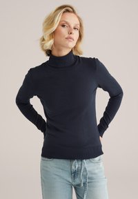 Tumši zila turtlenecka džemperis, izgatavots no mīksta adījuma, ar garām piedurknēm, ribinātām aprocēm un apakšmalu, apvienots ar gaismas mazgājumu džinsiem.