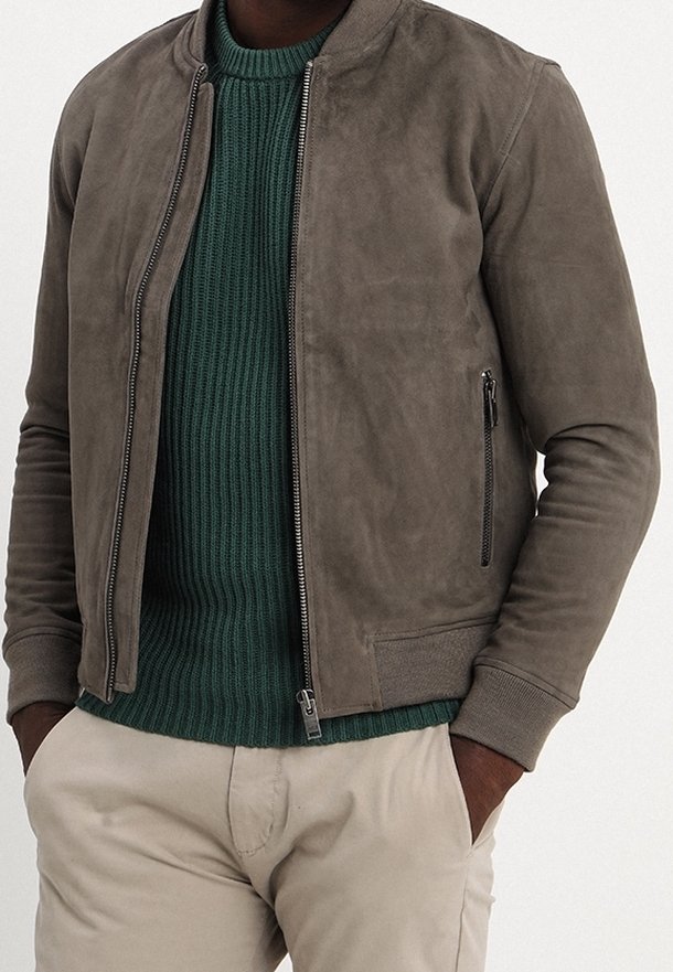 Giacca bomber in suede grigio attenuato con zip frontale, polsini a costine e due tasche laterali. Indossata sopra un maglione verde testurizzato.