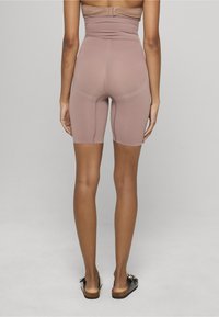 Kobieta w dopasowanych szortach w kolorze mauve z wysokim stanem i czarnych butach typu slip-on, stoi tyłem do kamery na jednolitym tle.