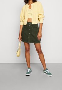 Žlutý žebrovaný kardigan, světle zelený crop top, tmavě zelená manšestrová sukně s předními knoflíky a zelené tenisky. Malá žlutá kabelka s akcenty.