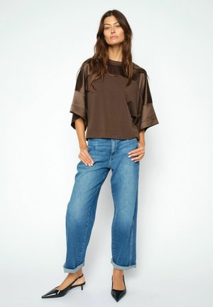 Donna in piedi su sfondo bianco con una maglia marrone larga, jeans blu con risvolto e décolleté slingback nere con punta.
