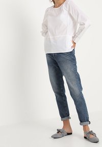 Camicia bianca a maniche lunghe con scollatura rotonda, abbinata a jeans a gamba larga di un blu chiaro. Accompagnata da scarpe slip-on a scacchi bianchi e neri.