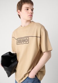Beige bomull T-skjorte med et svart brodert logo foran. Korte ermer og avslappet passform. Kombinert med en svart syntetisk lærveske.