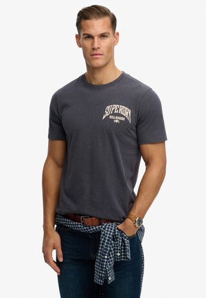 Camiseta de algodón de color gris oscuro con un logo en la parte frontal, mangas cortas y escote de tripulación. Llevada con jeans azules y una camisa a cuadros atada a la cintura.