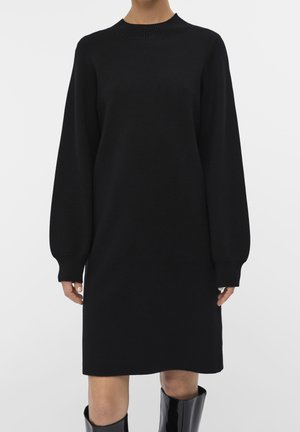 Robe noire en tricot avec un col montant et des manches longues, munie de poignets côtelés. Le modèle est droit et s'étend jusqu'à mi-cuisse.