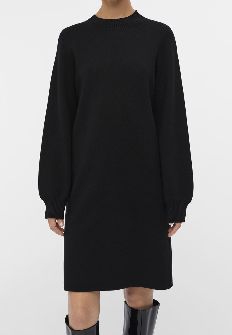 Robe noire en tricot avec un col montant et des manches longues, munie de poignets côtelés. Le modèle est droit et s'étend jusqu'à mi-cuisse.