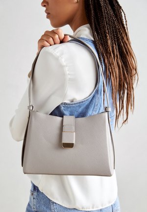 Femme aux cheveux tressés portant un chemisier blanc et un gilet en denim, tenant un sac à main structuré gris clair sur l'épaule.