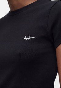 Musta ribbine lyhythihainen paita, jossa valkoinen "Pepe Jeans" kirjailtu vasemmassa rinnassa, henkilön päällä.