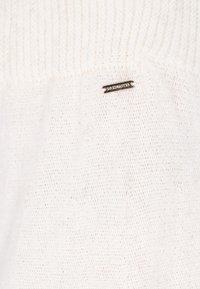 Pull en tricot crème clair avec une texture côtelée et une étiquette logo en métal. Le tissu semble doux et a une fine trame ouverte.