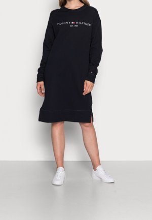 Nainen, joka pukee mustan Tommy Hilfiger -college-paidan pitkähihaista mekkoa logolla ja valkoiset lenkkarit, seisoo harmaalla lattialla valkoista seinää vasten.
