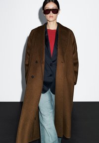 Brązowy podwójnie zapinany płaszcz na niebieskiej marynarce, czerwony sweter z okrągłym dekoltem i jasnoniebieskie dżinsy. Model w dużych ciemnych okularach przeciwsłonecznych.