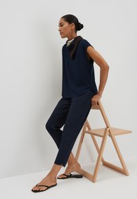 Haut sans manches bleu marine, pantalons sombres ajustés et tongs noires. Une écharpe à motifs dans des nuances de marron et de vert est portée autour du cou.