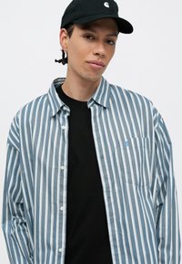Pruhovaná modro-biela košeľa s gombíkmi, má vrecko na hrudi a malé logo, nosená na čiernom tričku, doplnená čiernou čiapkou.