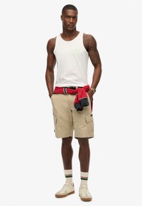 Top sin mangas blanco con un logo verde, combinado con pantalones cortos de cargo beige. Sudadera roja atada alrededor de la cintura y zapatillas color crema con rayas verde-rojas.