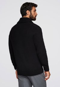 Czarny sweter z teksturowym splotem, z szerokim kołnierzem i ściągaczami na mankietach. Noszony na szarych spodniach, widok z tyłu.