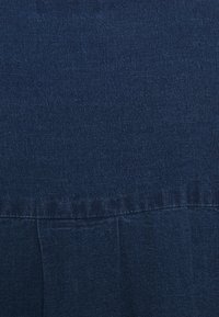 Tissu en denim bleu foncé, présentant une texture lisse avec des variations subtiles. Comprend des coutures verticales et un détail de ourlet froncé.