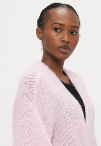 Ροζ πλεκτή ζακέτα με ανοιχτό μπροστινό μέρος, υφή νήματος και άνετη εφαρμογή, με βαθύ ντεκολτέ V και φαρδιά μανίκια.