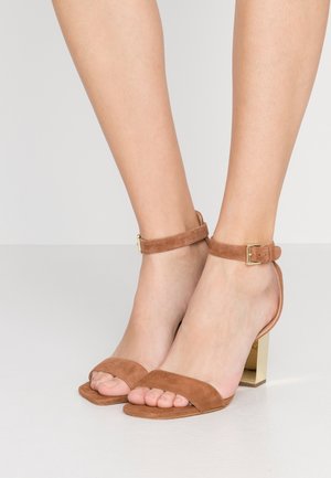 Pieds chaussés de sandales à talons hauts en daim marron, à bout ouvert, avec des brides autour de la cheville et des talons blocs dorés sur un fond clair uni.