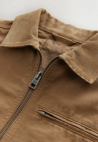 Giacca in velluto a coste marrone con zip frontale, colletto pieghevole e tasca sul petto. Presenta un tessuto testurizzato e una cerniera argentata.