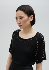 Černé tričko s krátkým rukávem, které má kovové nýty kolem výstřihu a rukávů, vyrobené z měkkého materiálu, se vyšitým detailem srdce.