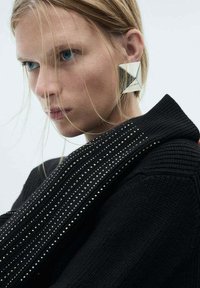 Schwarzer Strickpullover mit strukturierten Streifen und silbernen Akzenten. Das Model trägt einen großen geometrischen silbernen Ohrring, der das moderne Design betont.