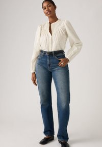 Γυναίκα που φοράει λευκό πουκάμισο με μακριά μανίκια, μπουσουλιασμένο σε ψηλόμεσο μπλε τζιν παντελόνι με μαύρη ζώνη και μαύρα παπούτσια, σε απλό φόντο.