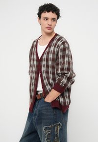 Cardigan od mekog, pahuljastog materijala u bordo i sivoj karo uzorku, s V-izrezom, gumbima i rebrastim manžetama, u kombinaciji s trapericama.