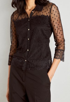 Czarna bluzka z przezroczystymi rękawami w groszki, koronkowymi kwiatowymi zdobieniami i złotymi guzikami. Posiada kołnierzyk oraz koronkowy brzeg z falbanką.
