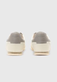 Sneaker in pelle crema e grigia con finitura testurizzata, suola piatta e una leggera personalizzazione sul tallone. Forma rotonda con dettagli di design minimalisti.