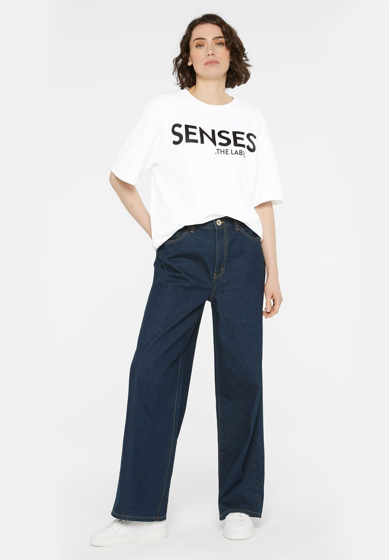 White t-shirt with black print "SENSES .THE LAB" paired with dark blue wide-leg denim jeans and white sneakers, showcasing a casual outfit.