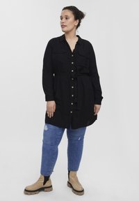 Vestido camisero negro con botones, mangas largas, lazo en la cintura y dos bolsillos en el pecho; combinado con jeans azules desgastados y botas de tobillo beige.