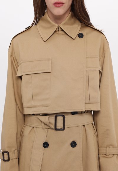 Trench coat beige a giacca corta con colletto abbottonato, doppie tasche sul petto e una cintura. Presenta una texture sottile e bottoni neri a contrasto.