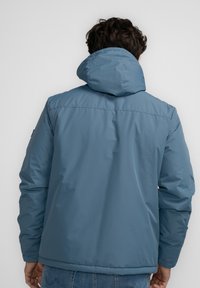 Chaqueta acolchada azul claro con capucha, con una textura suave y puños elásticos. La vista trasera muestra detalles de costura y un ajuste relajado.
