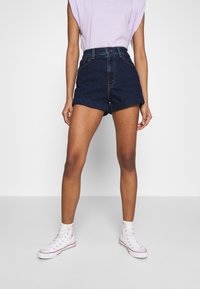 Denim shorts in dark blue with a high waist, rolled cuffs, and front pockets, paired with white high-top sneakers and a lilac sleeveless top.