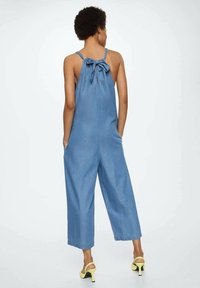 Combinaison en denim bleu avec jambes larges, bretelles réglables à nouer dans le dos et deux poches latérales. Présente une coupe décontractée et une texture douce.