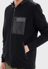 Felpa nera con zip e finitura texturizzata, caratterizzata da una tasca frontale nera e due tasche laterali con zip. Il cappuccio è realizzato in materiale liscio.