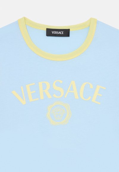 Ljusblå Versace t-shirt med gul krage, gul "VERSACE" text och Medusa-huvud logo centrerad under texten.