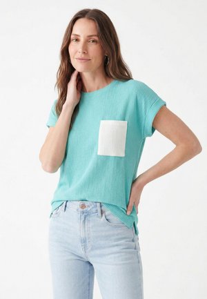 Kvinde med langt brunt hår iført en turkis ribbet kortærmet top med en hvid lomme og lyseblå jeans, poserer med den ene hånd på hoften.