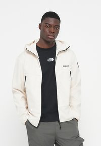 Lys cremefarvet softshell-jakke med lynlås, kontrasterende sorte detaljer og hætte. Sort logo på brystet, stylet med mørk bluse og cargo-bukser.