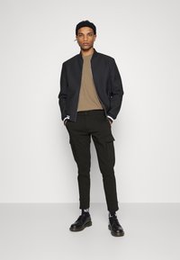 Sort bomberjakke med hvide stribede manschetter, brun t-shirt, olivengrøn cargo-buks og sorte sko. Sættet har et stilrent, moderne design.