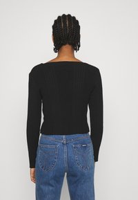 Haut noir côtelé à manches longues avec un col large, associé à un jean bleu avec des coutures visibles et des détails de poche arrière.