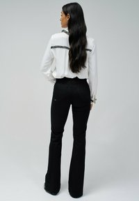 Blusa blanca de manga larga con ribetes negros, combinada con jeans acampanados negros ajustados, vista desde la parte posterior, con el cabello cayendo en cascada.