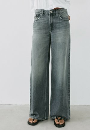 Person iført falmede grå jeans med vide ben parret med sorte flip-flops, stående på en lysegrå gulv mod en hvid væg.