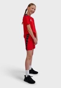 Jeune fille souriante aux cheveux tressés portant une tenue de sport rouge et des baskets noires, debout devant un fond uni.