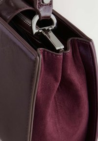 Sac à main bordeaux avec un extérieur en cuir brillant et un intérieur en daim doux. Équipé de quincaillerie en métal argenté et d'une fermeture éclair avec un tirette en métal.