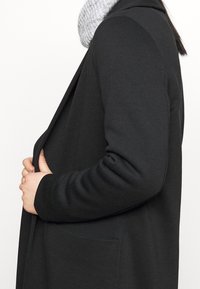 Schwarzer langer Mantel mit einer lässigen Silhouette, ausgestattet mit einer Vordertasche und einem weichen grauen Strickschal. Glatte Textur und schlichtes Design.