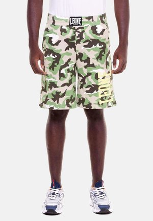 Uomo che indossa pantaloncini mimetici verdi, beige e marroni con logo "LEONE" in vita e sulla gamba sinistra, abbinati a scarpe da ginnastica bianche e blu navy.