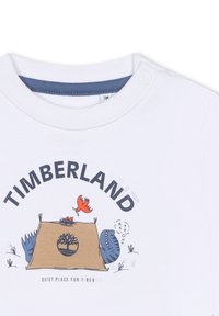 T-shirt bianca in cotone con colletto blu, stampa grafica di una tenda con un dinosauro e un uccello, e testo che recita "TIMBERLAND."