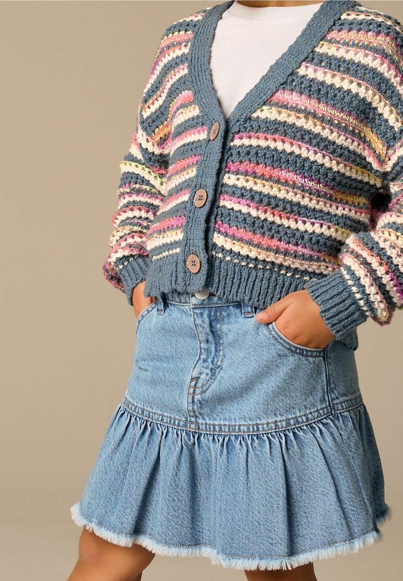 Enfant portant un cardigan tricoté rayé multicolore avec des boutons en bois sur une chemise blanche et une jupe en jean bleu clair à volants.