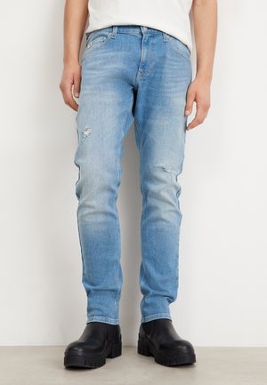 Mężczyzna w jasnoniebieskich obcisłych jeansach z małymi rozdarciami na udach i czarnych masywnych butach, stojący na białym tle.